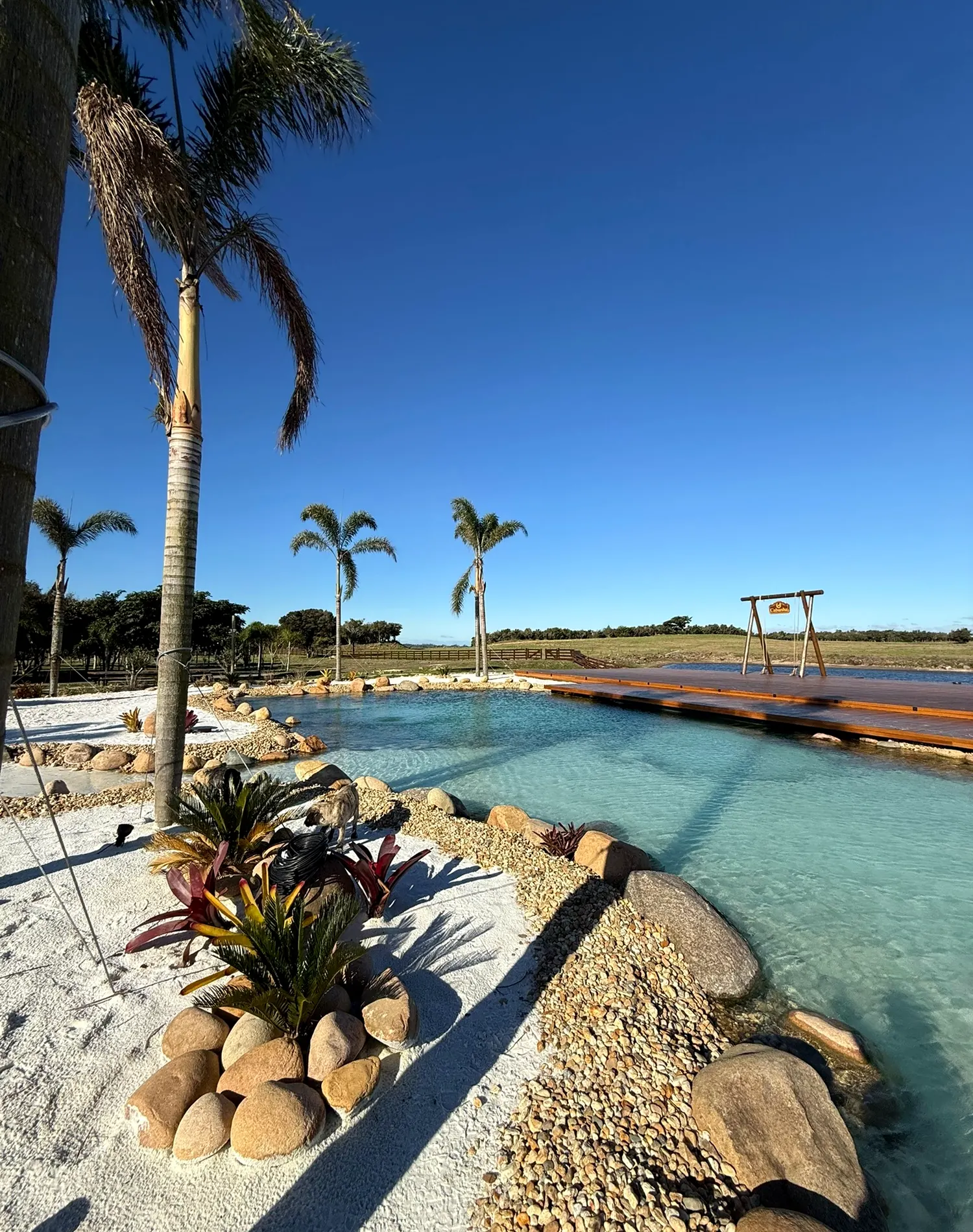 Piscina natural com água cristalina, areia branca, rochas e deck de madeira
